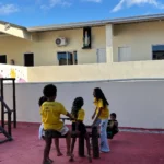 Quatro crianças brincam em um parquinho com piso pintado de vermelho, localizado na área externa de um prédio de dois andares com paredes amarelas e brancas. Três meninas vestem camisetas amarelas e calças ou shorts escuros, enquanto um menino está sentado na ponta oposta do brinquedo de madeira escura.