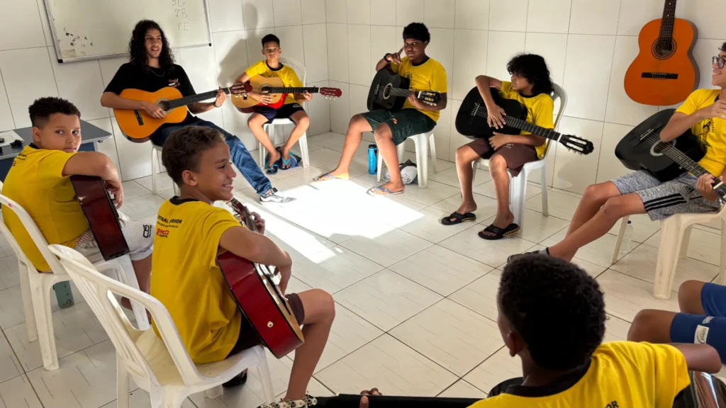 Sete crianças e um jovem instrutor estão sentados em círculo, em cadeiras de plástico brancas, tocando violão. O instrutor veste uma camisa preta e calça jeans, enquanto os alunos usam camisas amarelas de uniforme.