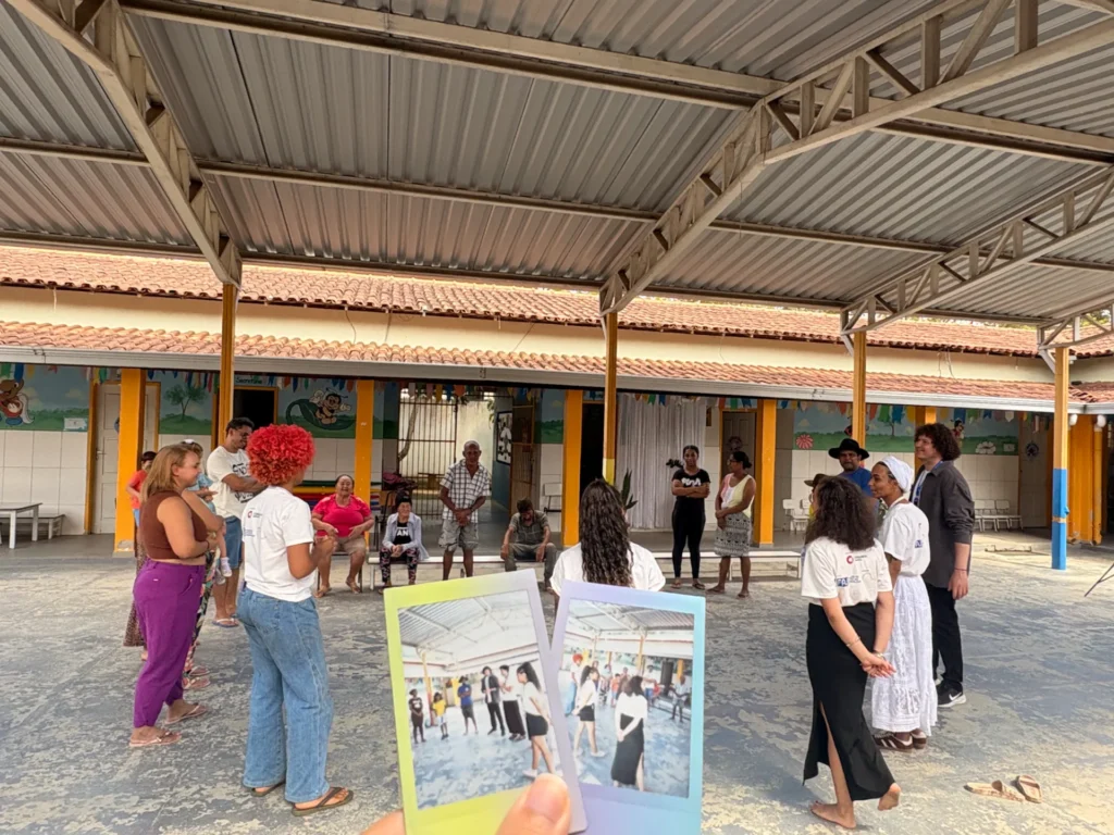 Duas fotos instantâneas estilo Polaroid são seguradas em primeiro plano, mostrando pessoas reunidas em um pátio.
Ao fundo, o mesmo grupo real de pessoas está disposto em círculo no próprio pátio coberto da creche.