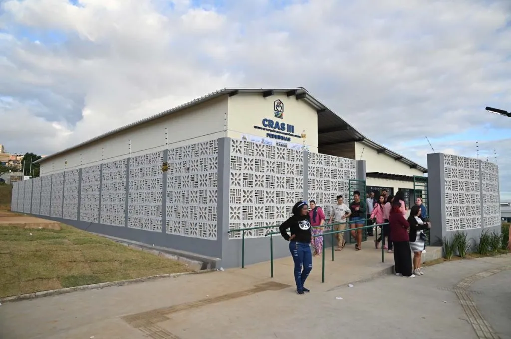Fachada do prédio do CRAS III Pedrinhas. Na frente do edifício, um grupo de pessoas desce uma rampa de acessibilidade com corrimão verde. Uma mulher de blusa preta e calça jeans destaca-se em pé no primeiro plano, à frente da rampa. O chão é de concreto cinza, há um gramado verde à esquerda e o céu ao fundo tem nuvens brancas.