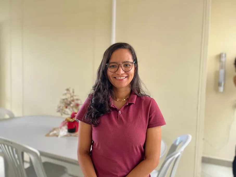 Uma mulher sorridente com pele parda, óculos de grau redondos e cabelos longos e ondulados castanhos está no centro. Ela veste uma camisa gola polo de cor vinho e um colar. Ao fundo, o cenário é de um ambiente interno com paredes beges claras, uma mesa cinza com cadeiras de plástico brancas e um pequeno enfeite natalino sobre a mesa.