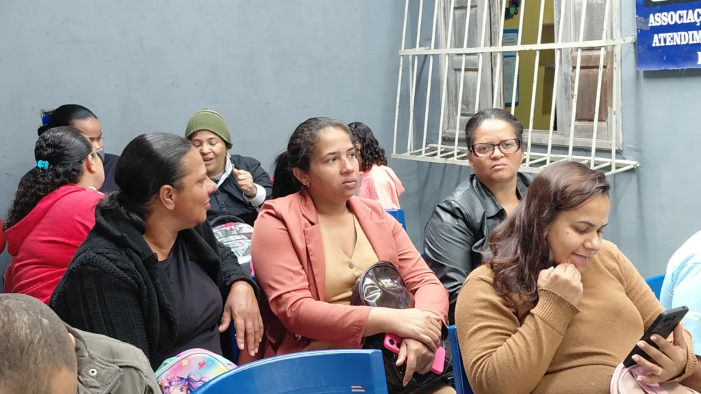 Fotografia colorida horizontal capturada em plano médio-longo mostra um grupo de cerca mulheres sentadas em cadeiras de plástico azuis em um local com paredes cinzas. Jéssica Machado, a mulher no centro, veste um blazer rosa seco sobre uma blusa bege.