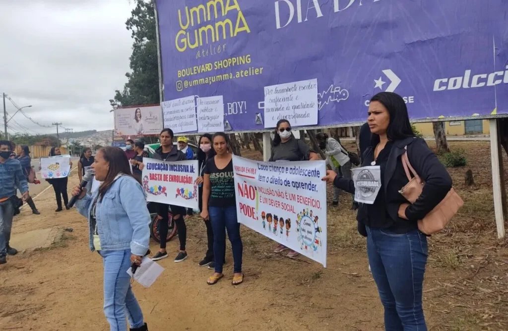 Fotografia colorida horizontal capturada em plano médio mostra um grupo de mulheres protestando ao ar livre em uma área de terra batida e asfalto, à frente de um grande outdoor roxo.