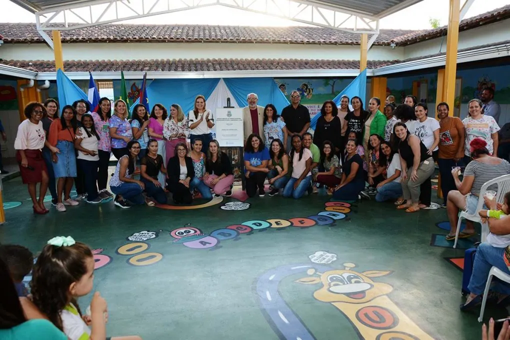 Um grande grupo de pessoas posa reunido no pátio coberto da creche do Senhorinha Cairo. Elas estão atrás de uma placa oficial de inauguração e diante de um chão verde decorado com desenhos coloridos.