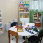 Uma mesa redonda branca com brinquedos coloridos ocupa o centro de uma sala de atendimento para crianças atípicas. Ao redor da mesa, há quatro cadeiras. o fundo e nas laterais, erguem-se armários e estantes brancas repletos de livros infantis, jogos em caixas coloridas, brinquedos e um globo terrestre. 
