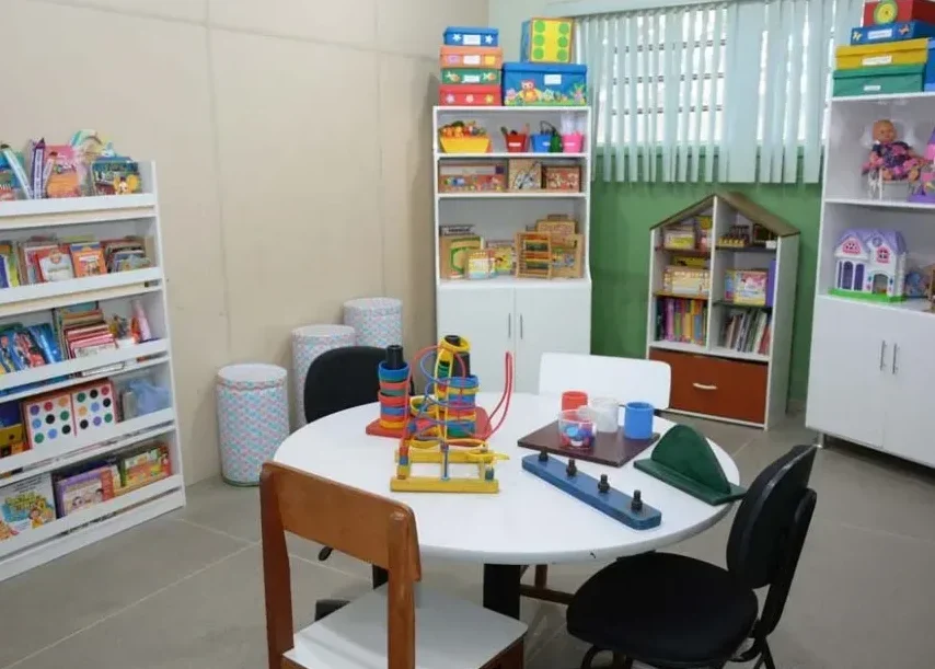 Uma mesa redonda branca com brinquedos coloridos ocupa o centro de uma sala de atendimento para crianças atípicas. Ao redor da mesa, há quatro cadeiras. o fundo e nas laterais, erguem-se armários e estantes brancas repletos de livros infantis, jogos em caixas coloridas, brinquedos e um globo terrestre. 