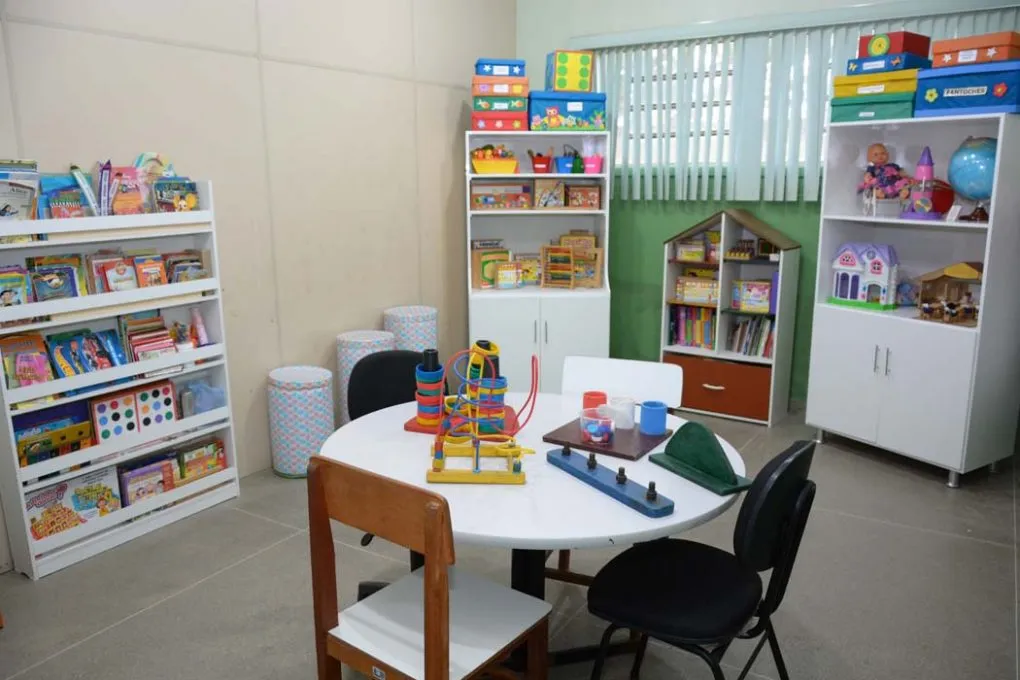 Fotografia colorida horizontal capturada em plano médio-longo mostra uma sala de Atendimento Educacional Especializado (AEE) organizada e sem pessoas. No centro, há uma mesa redonda branca cercada por cadeiras infantis e com vários brinquedos pedagógicos sobre ela, como um aramado sinuoso. Ao fundo, as paredes são divididas entre bege e verde, com uma janela de persianas verticais brancas.