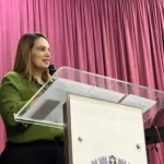 Prefeita Sheila Lemos veste uma blusa verde e uma calça preta. Ela segura um microfone com as mãos enquanto está em pé em um púlpito, que contém a logomarca da Prefeitura de Vitória da Conquista.