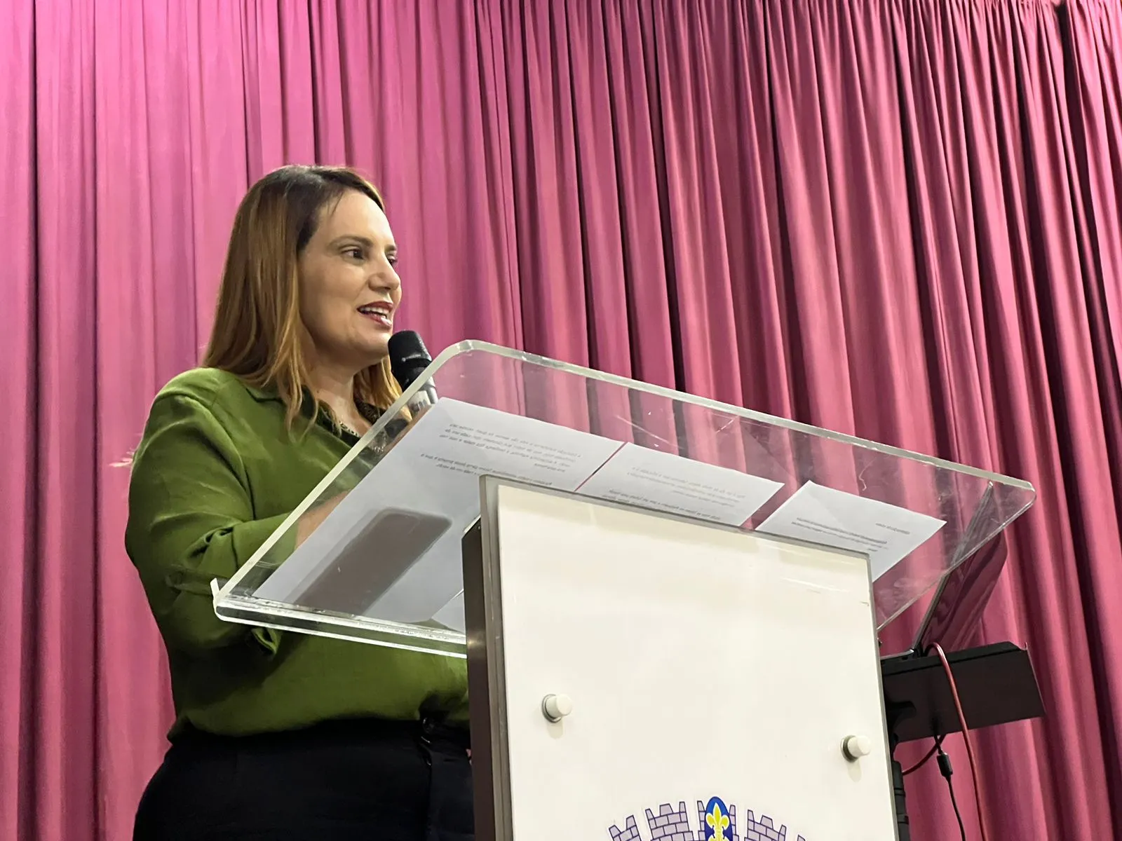 Prefeita Sheila Lemos veste uma blusa verde e uma calça preta. Ela segura um microfone com as mãos enquanto está em pé em um púlpito, que contém a logomarca da Prefeitura de Vitória da Conquista.