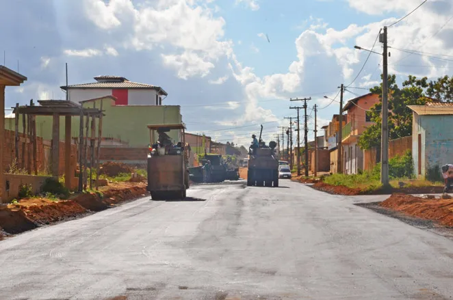 Máquinas de pavimentação asfáltica trabalham em uma rua de terra recém-asfaltada. Ao longo da via, há casas simples, postes de energia e terra vermelha nas laterais. O céu ao fundo está claro, com nuvens brancas e iluminadas pelo sol.